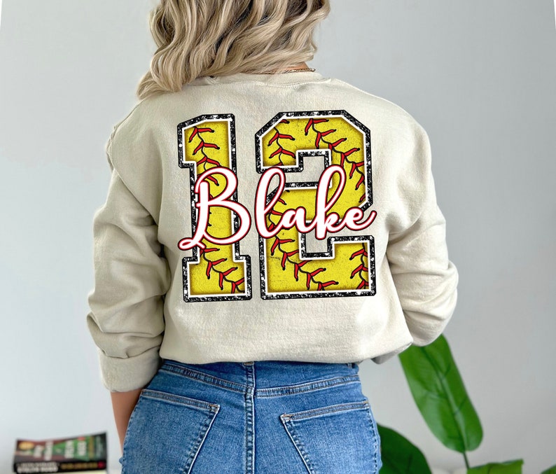 May include: A beige sweatshirt with a large number "12" printed on the back. The number is made up of two softball designs with red stitching and a black outline. The name "Blake" is written in pink cursive inside the number.