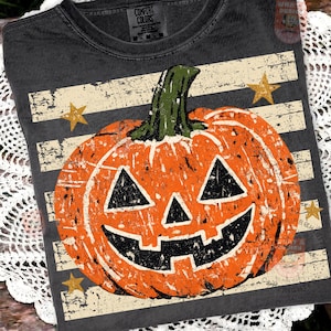 May include: A charcoal gray t-shirt featuring a distressed graphic of an orange jack-o'-lantern with a green stem. The design includes horizontal white stripes and gold stars. The neck label reads Comfort Colors, and the fabric has a vintage wash.