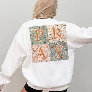 May include: A white long-sleeve sweatshirt featuring a large square graphic on the back. The graphic is divided into four quadrants with floral patterns in shades of sage green, cream, and orange, displaying the orange text P R A Y across them.