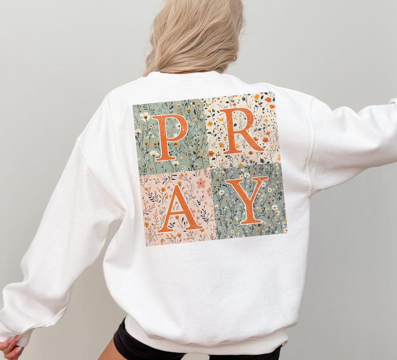 May include: A white long-sleeve sweatshirt featuring a large square graphic on the back. The graphic is divided into four quadrants with floral patterns in shades of sage green, cream, and orange, displaying the orange text P R A Y across them.
