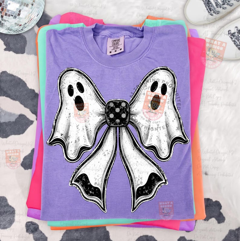 Op de afbeelding: Een lavendelkleurig T-shirt met korte mouwen en een grote afbeelding van een strik gemaakt van twee witte geesten met zwarte ogen en monden. Het midden van de strik is een zwarte dobbelsteen met witte stippen. Het shirt heeft een ronde hals.