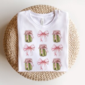 Op de afbeelding: Wit T-shirt met een herhalend patroon van roze strikken en potten met groene augurken.