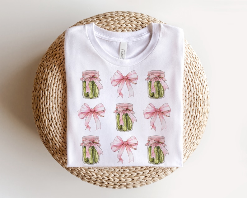 Op de afbeelding: Wit T-shirt met een herhalend patroon van roze strikken en potten met groene augurken.