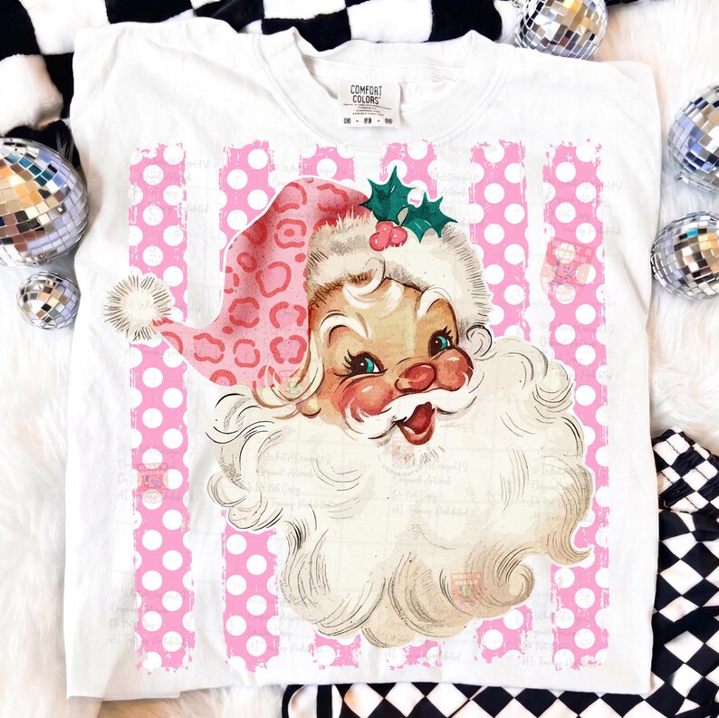 Pode incluir: Uma t-shirt branca com um gr&aacute;fico de um Pai Natal de estilo vintage a usar um gorro cor-de-rosa com padr&atilde;o de leopardo, um pompom branco e folhas de azevinho verdes. O fundo do gr&aacute;fico consiste em riscas verticais cor-de-rosa com bolas.