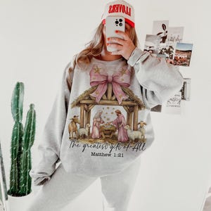 May include: A gray sweatshirt with a pink bow and a nativity scene graphic. The text "The greatest gift of all" and "Matthew 1:21" are printed below the image.