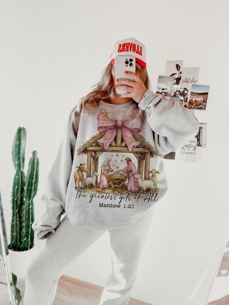 May include: A gray sweatshirt with a pink bow and a nativity scene graphic. The text "The greatest gift of all" and "Matthew 1:21" are printed below the image.