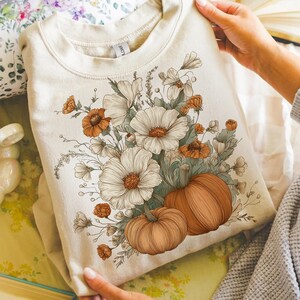 Pu&ograve; includere: Felpa color crema con un design floreale e zucche. La grafica include grandi fiori bianchi, accenti arancioni e due zucche. Il design &egrave; centrato sulla parte anteriore della felpa, creando un tema autunnale.