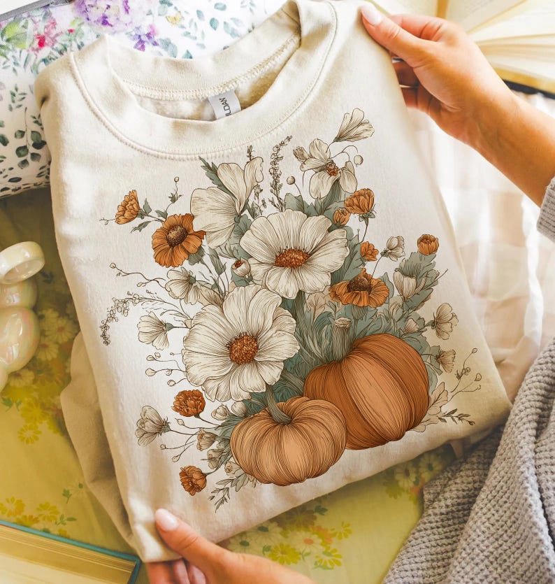 Pu&ograve; includere: Felpa color crema con un design floreale e zucche. La grafica include grandi fiori bianchi, accenti arancioni e due zucche. Il design &egrave; centrato sulla parte anteriore della felpa, creando un tema autunnale.