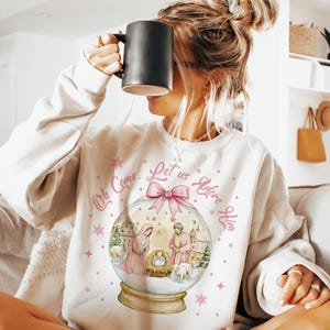 K&ouml;nnte beinhalten: Ein wei&szlig;er Pullover mit einem rosa-wei&szlig;en Schneekugel-Design. Die Schneekugel zeigt eine Weihnachtskrippe mit Maria, Josef und dem Jesuskind. Der Text um die Schneekugel lautet "Oh Come, Let Us Adore Him".