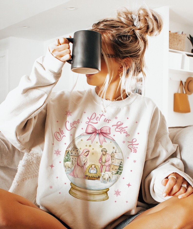 K&ouml;nnte beinhalten: Ein wei&szlig;er Pullover mit einem rosa-wei&szlig;en Schneekugel-Design. Die Schneekugel zeigt eine Weihnachtskrippe mit Maria, Josef und dem Jesuskind. Der Text um die Schneekugel lautet "Oh Come, Let Us Adore Him".