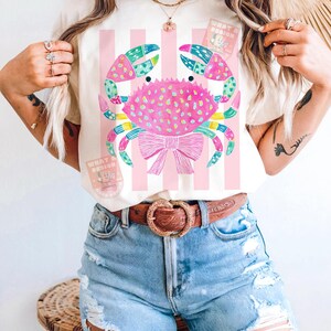 May include: Cream-colored t-shirt featuring a colorful crab design with a pink bow and vertical pink stripes. The crab has blue, green, and pink accents. The shirt also has the text "WHAT A DESIGN" on it.