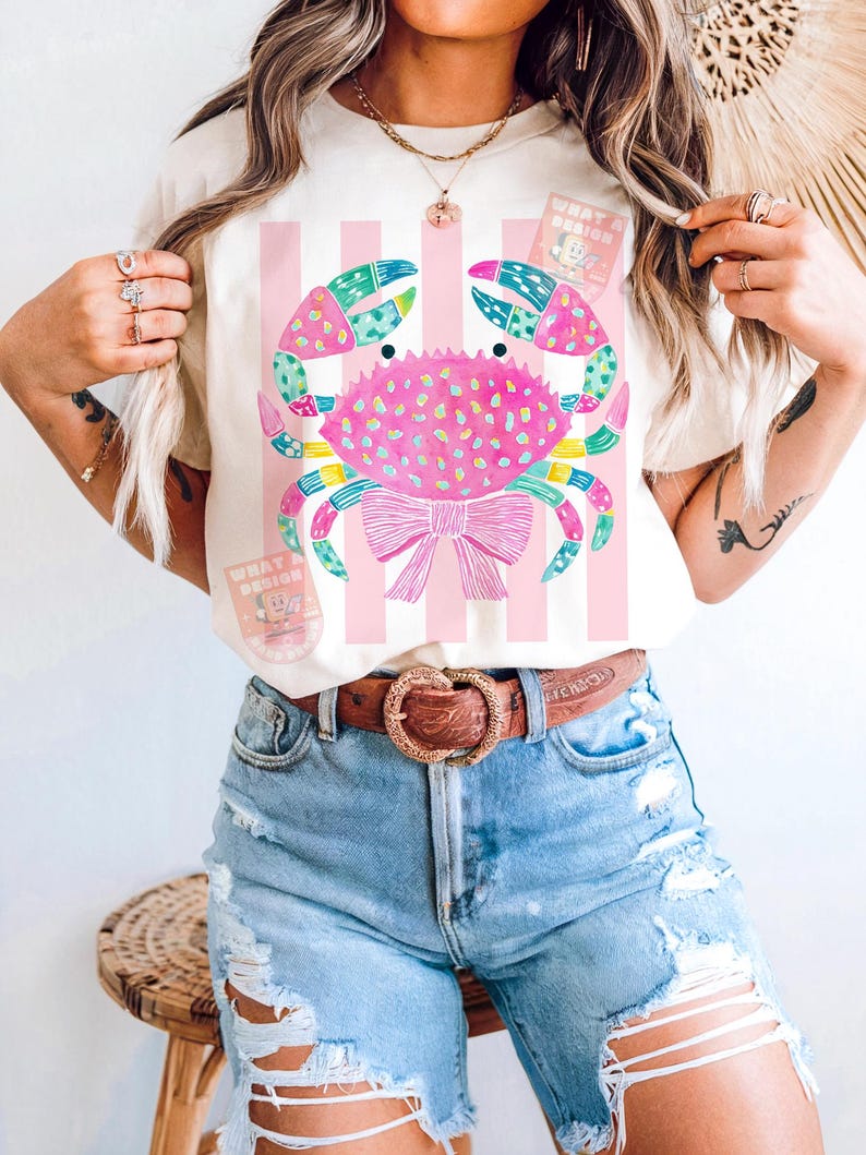 May include: Cream-colored t-shirt featuring a colorful crab design with a pink bow and vertical pink stripes. The crab has blue, green, and pink accents. The shirt also has the text "WHAT A DESIGN" on it.