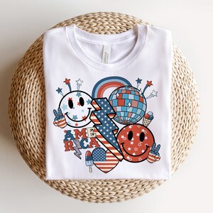 May include: White t-shirt with a red, white and blue graphic design featuring a smiley face, a disco ball, and the word "AMERICA".