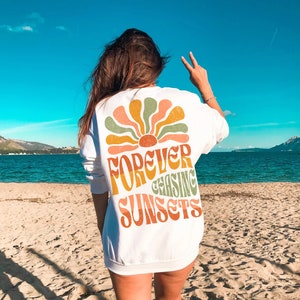 May include: White sweatshirt with a retro-style graphic on the back. The design features a stylized sunburst in orange, green, and peach, with the words "FOREVER CHASING SUNSETS" in a matching font. The sweatshirt is worn on a beach.