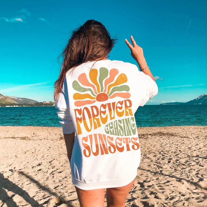 May include: White sweatshirt with a retro-style graphic on the back. The design features a stylized sunburst in orange, green, and peach, with the words "FOREVER CHASING SUNSETS" in a matching font. The sweatshirt is worn on a beach.