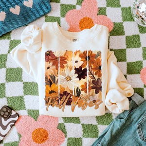 May include: Cream-colored sweatshirt with a floral design in shades of brown, orange, and white, with the words "Fall Vibes" in a cursive font. The sweatshirt is laid on a green and white checkered rug with pink flower accents, next to blue jeans.