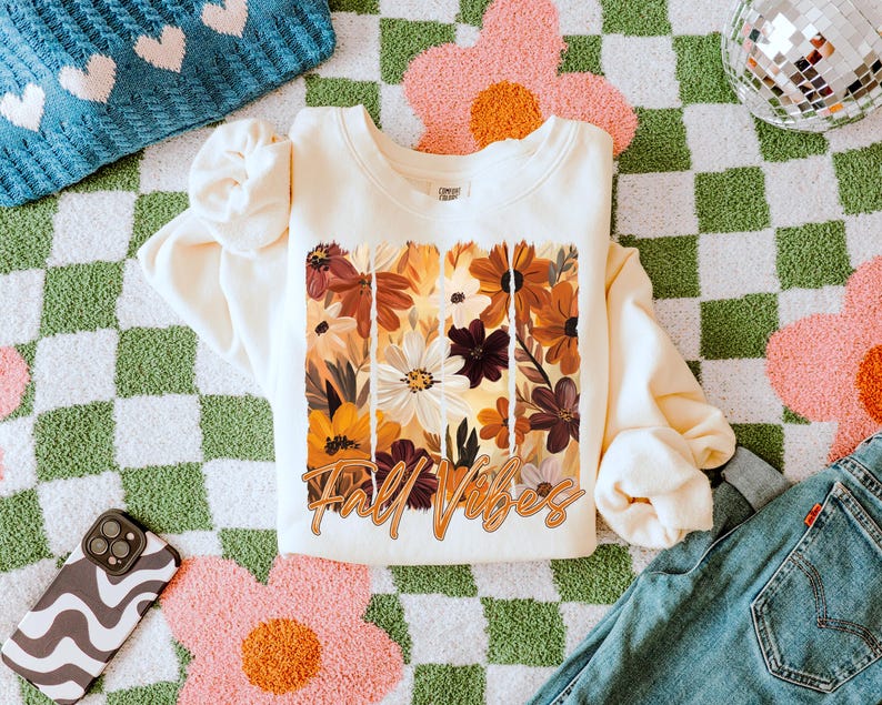 May include: Cream-colored sweatshirt with a floral design in shades of brown, orange, and white, with the words "Fall Vibes" in a cursive font. The sweatshirt is laid on a green and white checkered rug with pink flower accents, next to blue jeans.