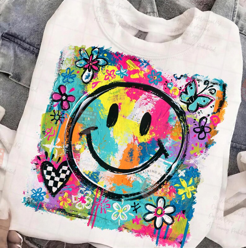 May include: White sweatshirt featuring a vibrant, colorful graphic. The design includes a large smiley face with a black outline, surrounded by flowers, butterflies, and a heart with a checkered pattern. The background is a mix of bright colors.