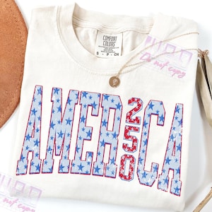 May include: Cream-colored t-shirt with the word "AMERICA" in blue stars and the number "250" in red and white stripes. A gold necklace with a pendant rests on the shirt. The shirt is a Comfort Colors brand, size S.