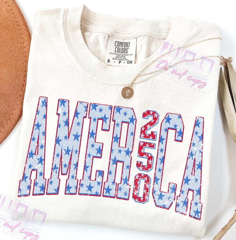 May include: Cream-colored t-shirt with the word "AMERICA" in blue stars and the number "250" in red and white stripes. A gold necklace with a pendant rests on the shirt. The shirt is a Comfort Colors brand, size S.