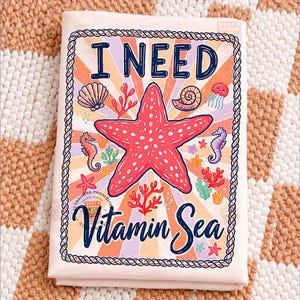 May include: A light pink beach towel with the text "I NEED Vitamin Sea" in navy blue script. The towel features a red starfish, seahorses, seashells, and coral illustrations. The design is framed with a rope border.