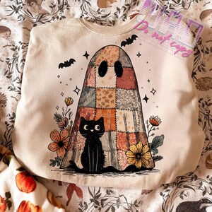 May include: Cream-colored sweatshirt featuring a patchwork ghost design with a black cat, flowers, and bats. The ghost has a patchwork design in shades of orange, brown, and beige. The black cat sits at the bottom of the ghost. Bats and stars are scattered around the ghost.