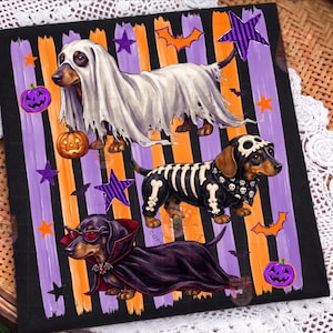 May include: A black fabric square featuring three dachshunds in Halloween costumes: one as a ghost, one as a skeleton, and one as a vampire. The background has vertical orange and purple brushstrokes, orange pumpkins, purple stars, and bats.