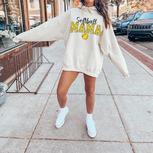 May include: A woman wearing a cream-colored hoodie with the text "Softball Mama" in black and yellow. The hoodie has a large pocket on the front and a drawstring hood. She is wearing white sneakers.