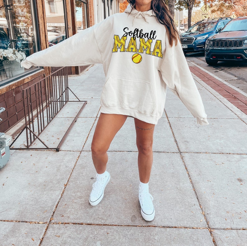 May include: A woman wearing a cream-colored hoodie with the text "Softball Mama" in black and yellow. The hoodie has a large pocket on the front and a drawstring hood. She is wearing white sneakers.