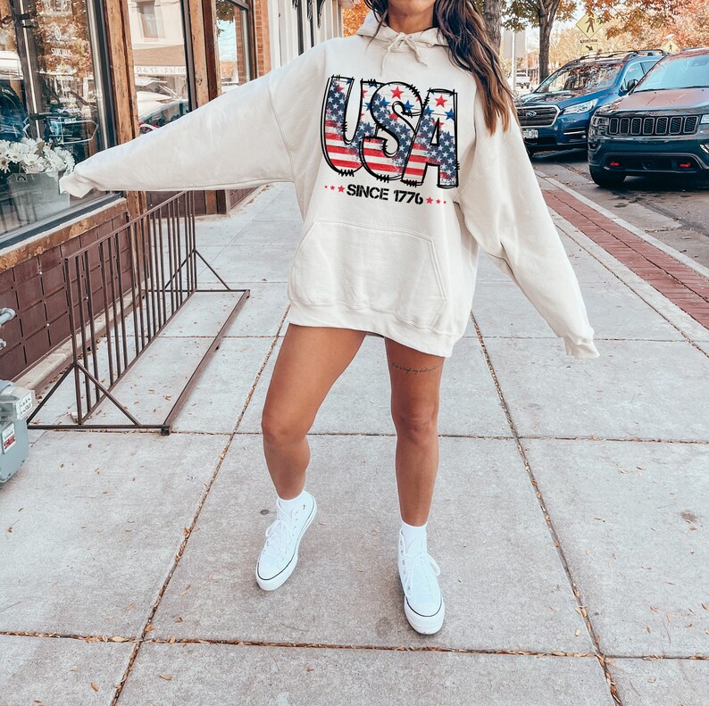 May include: A cream-colored hoodie with a graphic of the word "USA" in red, white, and blue with stars. The hoodie has a drawstring hood and a kangaroo pocket. The text "Since 1776" is printed below the graphic.