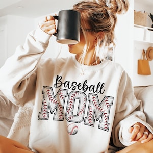 May include: White sweatshirt with the text "Baseball Mom" in black lettering. The letters are outlined in red and white stitching, resembling a baseball. A baseball is below the text.