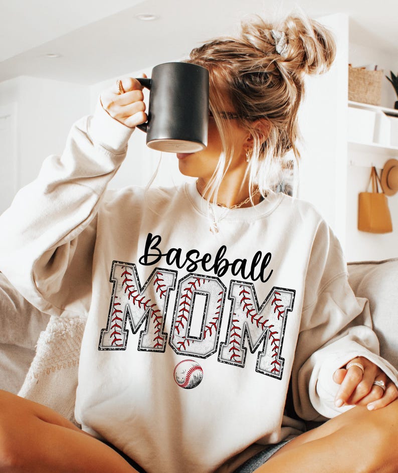 May include: White sweatshirt with the text "Baseball Mom" in black lettering. The letters are outlined in red and white stitching, resembling a baseball. A baseball is below the text.
