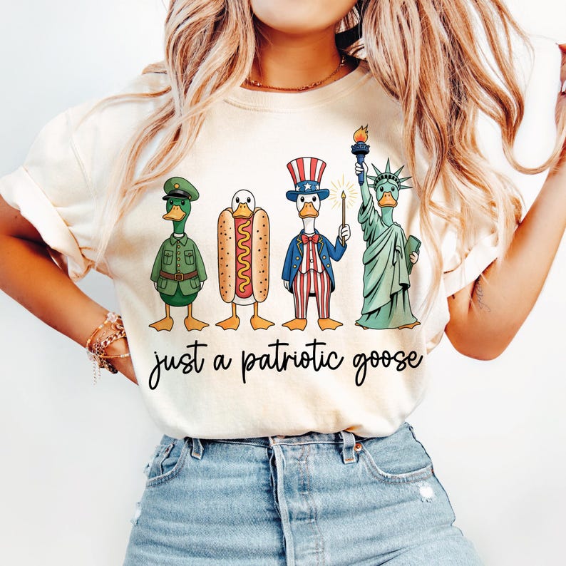 May include: Cream-colored t-shirt featuring a whimsical illustration of patriotic ducks. The ducks are dressed as a soldier, a hot dog, Uncle Sam, and the Statue of Liberty. The text "just a patriotic goose" is written below the ducks.