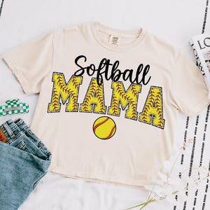 May include: A beige t-shirt with the text "Softball Mama" in black lettering with a yellow softball graphic. The text is outlined in brown stitching and the softball has red stitching.