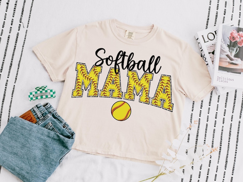 May include: A beige t-shirt with the text "Softball Mama" in black lettering with a yellow softball graphic. The text is outlined in brown stitching and the softball has red stitching.