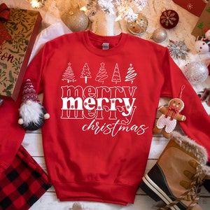 May include: A red sweatshirt with the words "merry merry christmas" in white, along with stylized Christmas tree graphics. The sweatshirt is surrounded by holiday decorations, including ornaments and gift boxes.