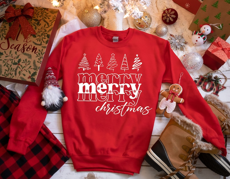 May include: A red sweatshirt with the words "merry merry christmas" in white, along with stylized Christmas tree graphics. The sweatshirt is surrounded by holiday decorations, including ornaments and gift boxes.