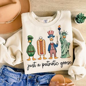 May include: Cream-colored sweatshirt with a graphic of four cartoon geese dressed as an army officer, a hot dog, Uncle Sam, and the Statue of Liberty. The text "just a patriotic goose" is written below the geese.