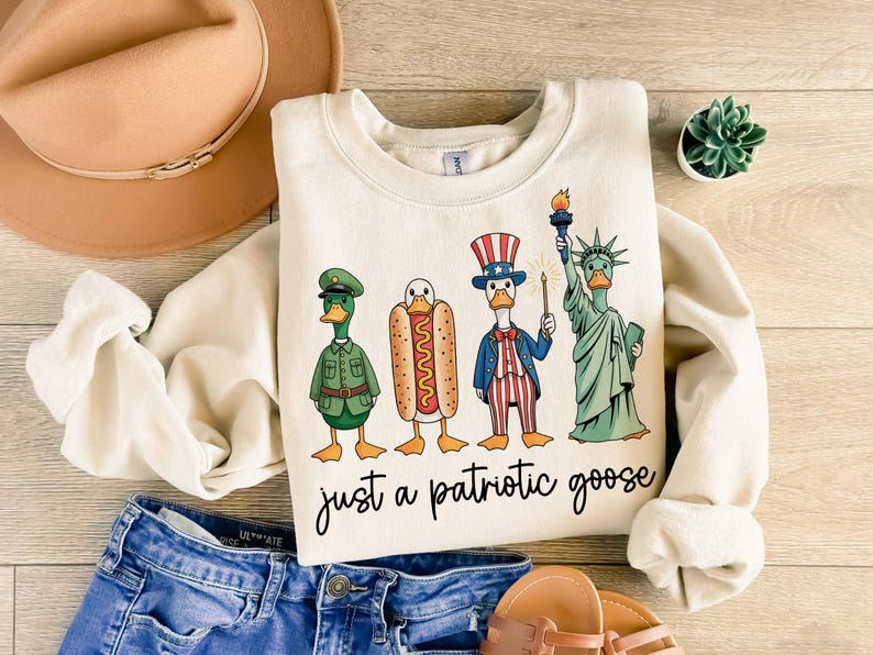 May include: Cream-colored sweatshirt with a graphic of four cartoon geese dressed as an army officer, a hot dog, Uncle Sam, and the Statue of Liberty. The text "just a patriotic goose" is written below the geese.