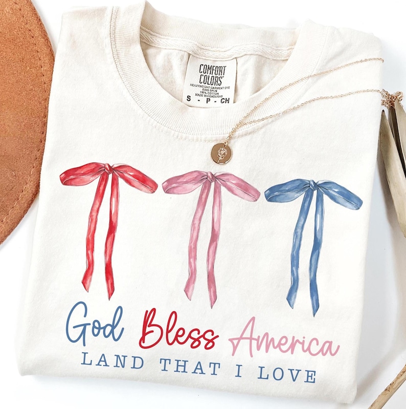 May include: A white t-shirt with a patriotic design featuring three red, white, and blue bows and the text "God Bless America Land That I Love".