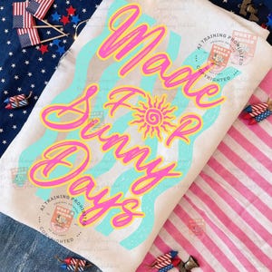 May include: A white t-shirt with the text "Made For Sunny Days" in pink and yellow, with a sun graphic. The design includes light blue wavy lines. The background features patriotic decorations.