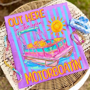 May include: A purple t-shirt with a colorful graphic of a pontoon boat, sun, and text that reads "OUT HERE in the open MOTORBOATIN'". The shirt is decorated with blue and white stripes. Accessories include a blue and white checkered hair clip and beaded necklaces.