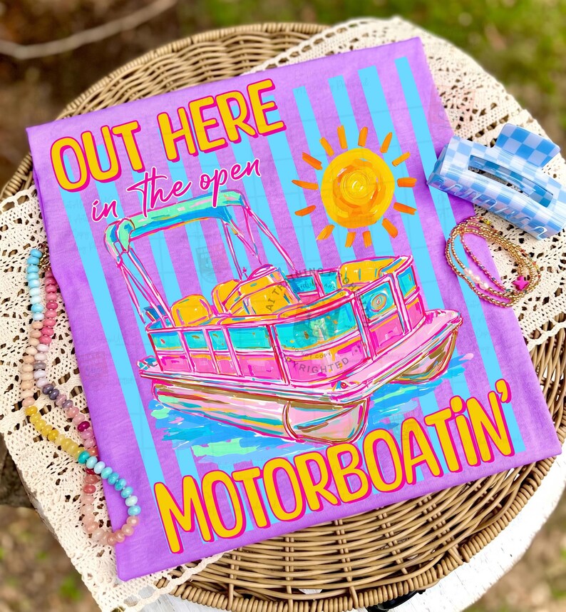 May include: A purple t-shirt with a colorful graphic of a pontoon boat, sun, and text that reads "OUT HERE in the open MOTORBOATIN'". The shirt is decorated with blue and white stripes. Accessories include a blue and white checkered hair clip and beaded necklaces.