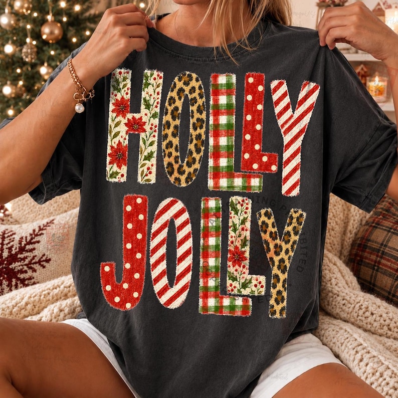 Op de afbeelding: Een antracietgrijs T-shirt met korte mouwen en de tekst HOLLY JOLLY in grote blokletters. Elke letter heeft een ander patroon, waaronder rode kerststerren, luipaardprint, rood-groene ruiten, rode stippen en rood-witte strepen.