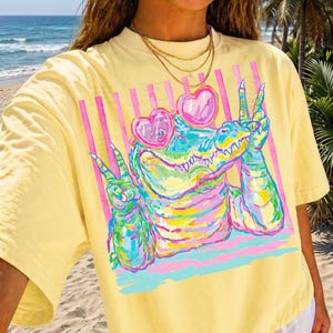 May include: Yellow t-shirt featuring a colorful alligator wearing heart-shaped sunglasses and making a peace sign. The graphic includes pink and blue stripes. The shirt is a relaxed fit.