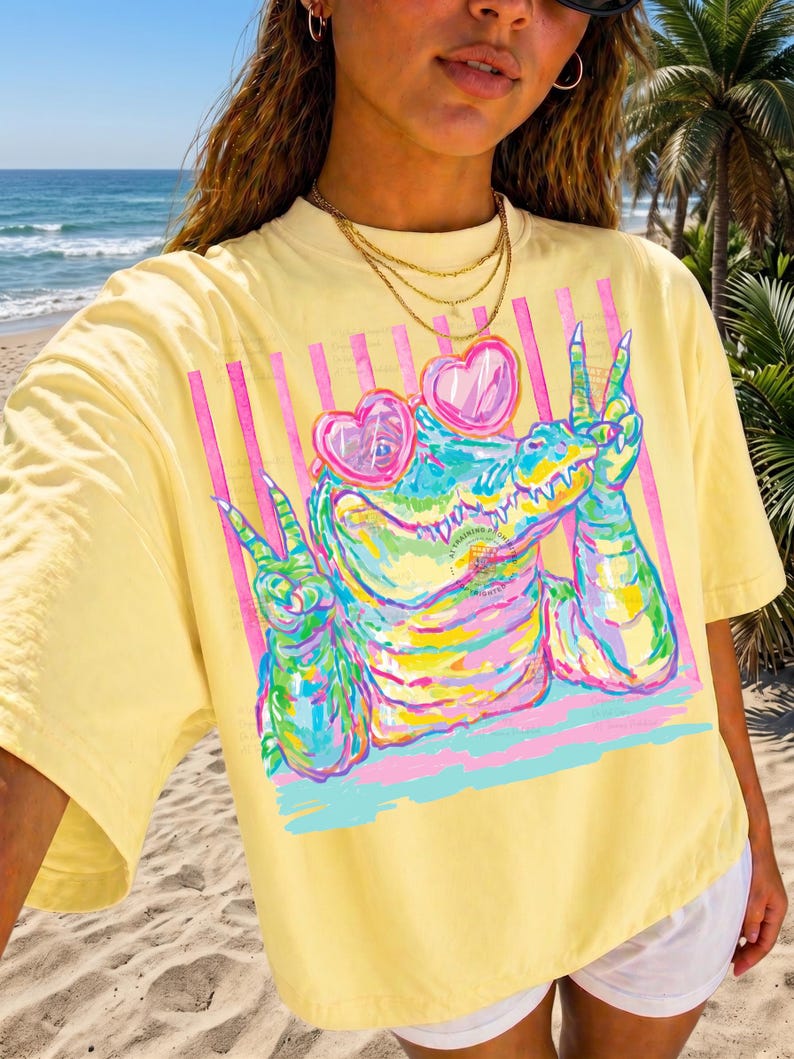 May include: Yellow t-shirt featuring a colorful alligator wearing heart-shaped sunglasses and making a peace sign. The graphic includes pink and blue stripes. The shirt is a relaxed fit.