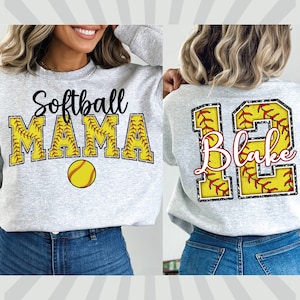 May include: Two gray sweatshirts with a softball theme. One sweatshirt has the text "Softball Mama" with a baseball graphic. The other sweatshirt has a large number "12" with the name "Blake" inside the number. Both sweatshirts have a glittery, baseball-stitched design.