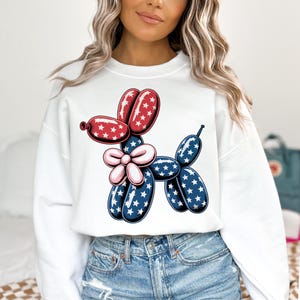 May include: White sweatshirt with a red, white, and blue balloon animal design. The balloon animal is shaped like a dog with a flower on its neck.