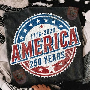 May include: Dark gray t-shirt featuring a distressed graphic celebrating America's 250th anniversary. The design includes a blue and red circular emblem with stars, the text "AMERICA," and the years "1776-2026" and "250 YEARS."
