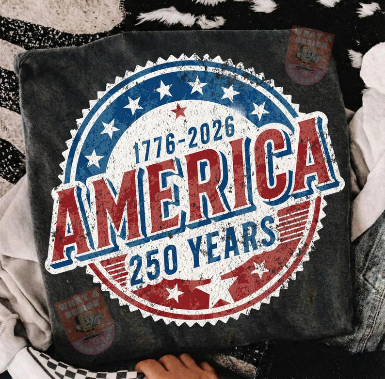 May include: Dark gray t-shirt featuring a distressed graphic celebrating America's 250th anniversary. The design includes a blue and red circular emblem with stars, the text "AMERICA," and the years "1776-2026" and "250 YEARS."